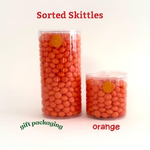 May include: Two clear plastic cylinders filled with orange Skittles candies. The text "Sorted Skittles" is at the top, with "gift packaging" and "orange" written below. Each cylinder has an orange sticker on the lid.