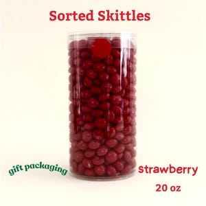 May include: A clear plastic cylinder filled with red strawberry Skittles. The text "Sorted Skittles" is at the top, with "strawberry" and "20 oz" at the bottom. The words "gift packaging" are in green on the left side.