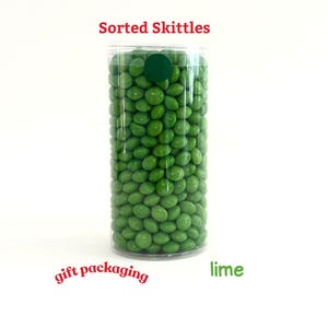 May include: A clear plastic tube filled with green Skittles candies. The text "Sorted Skittles" is at the top, with "gift packaging" and "lime" written below. A green sticker seals the top.