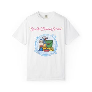 以下が含まれることがあります： 白いTシャツ。中央には「Sparkle Cleaning Service」というピンクの文字があり、その下に緑色のバケツ、スプレーボトル、黄色の手袋、青い布などの掃除用具が描かれた大きな水色のシャボン玉のグラフィックがプリントされています。