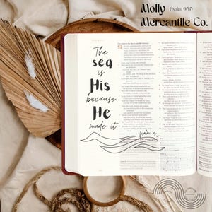 May include: An open book with the text "The sea is His because He made it" in black script. The book is on a neutral-toned surface with decorative elements, including dried leaves and feathers. The text "Molly Mercantile Co." is at the top.