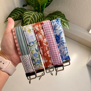 May include: A collection of five fabric wrist keychains with various patterns. The patterns include floral, gingham, and patchwork designs in colors like blue, pink, and orange. Each keychain has a metal clasp for attaching keys.