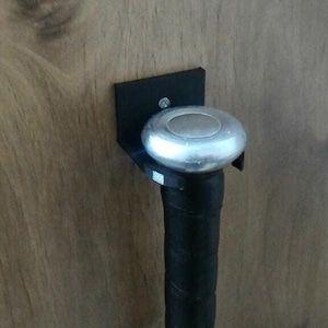May include: A black metal baseball bat holder mounted on a wooden surface. The holder has a black bracket and a silver-colored circular rest for the bat's end. The bat's black grip is visible.