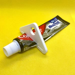 May include: A white plastic toothpaste tube squeezer rests on a tube of Colgate Max Fresh Charcoal Mint toothpaste. The toothpaste tube is dark gray with white text and red accents. The background is a bright yellow.
