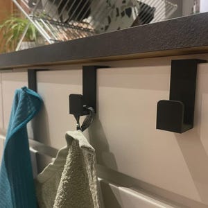 May include: Black metal over-the-door hooks. Two hooks are visible, hanging on a white cabinet door. A teal and a beige towel are hanging from the hooks. The hooks are designed to fit over the top of a cabinet door.