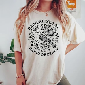 Pode incluir: Camiseta creme com um desenho gráfico preto de um pássaro empoleirado em um galho frondoso, rodeado por flores. O texto "RADICALIZED BY BASIC DECENCY" é impresso ao redor do desenho do pássaro. A camiseta tem mangas curtas.