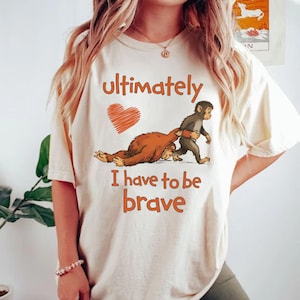 Op de afbeelding: Crèmekleurig T-shirt met de tekst "Ultimately I have to be brave" in oranje. De afbeelding toont een bruin wezen dat door een kleine grijze aap wordt getrokken, met een oranje hart erboven.