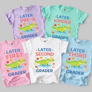 Könnte beinhalten: Fünf farbenfrohe Kinder-T-Shirts mit Schulmotiven. Jedes Shirt zeigt ein Cartoon-Krokodil, einen Apfel und Text, der die Klassenstufe angibt: Pre-K, Kinder, First, Second und Third Grader. Die Shirts sind in Lila, Grün, Rosa, Weiß und Blau gehalten.