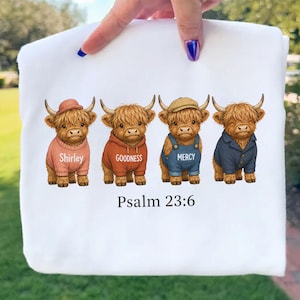 Könnte beinhalten: Weißes Sweatshirt mit einem Aufdruck von vier Highland-Rindern, die verschiedene Outfits tragen und mit Wörtern wie "Shirley" und "Goodness" beschriftet sind. Der Text "Psalm 23:6" ist unter den Kühen aufgedruckt.