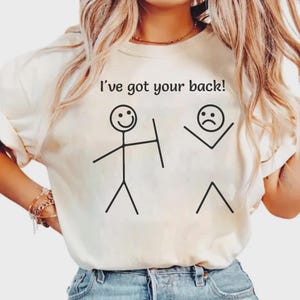 Può includere: T-shirt color crema con la frase "I've got your back!" e illustrazioni di omini stilizzati. Un omino ha una faccia sorridente e uno scudo, l'altro una faccia triste. La maglietta è un capo casual per tutti i giorni.