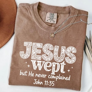 May include: A brown Comfort Colors t-shirt with the text "JESUS wept but He never complained John 11:35" in white. A silver necklace with a small pendant is draped across the shirt.