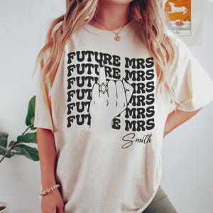 May include: A cream-coloured t-shirt featuring the text "FUTURE MRS" stacked vertically, with "Smith" written below. A hand with a ring on the ring finger is in the centre. The shirt is a casual style.