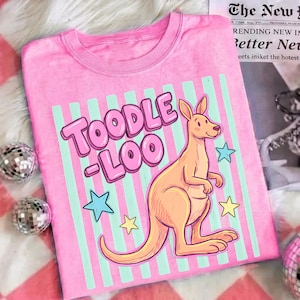 Op de afbeelding: Roze t-shirt met de tekst "Toodle Loo" in bubbelletters, een cartoon kangoeroe en sterrenafbeeldingen. Het shirt heeft een ronde hals en een lichtroze achtergrond met verticale strepen.