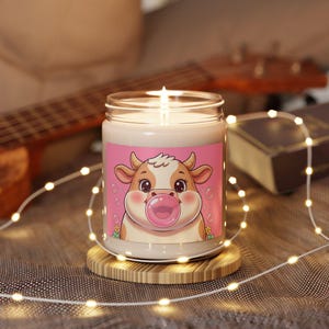 Könnte beinhalten: Eine brennende Kerze in einem Glas mit einem rosa Etikett, das eine Cartoon-Kuh zeigt, die eine Kaugummiblase bläst. Die Kerze steht auf einem runden Holzuntersetzer, mit Lichterketten und einer Gitarre im Hintergrund.
