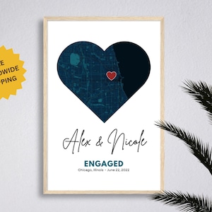 May include: A framed print with a navy blue heart-shaped map and a red heart marker. The names "Alex & Nicole" are written in cursive, with "ENGAGED" and the date "June 22, 2022" below. A yellow starburst graphic is in the upper left corner.