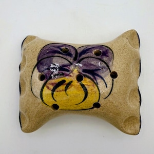 May include: A rectangular ceramic flower frog with a beige speckled finish and scalloped edges. The center features a hand-painted purple and yellow pansy design. The piece contains six circular holes distributed across the surface for stems.