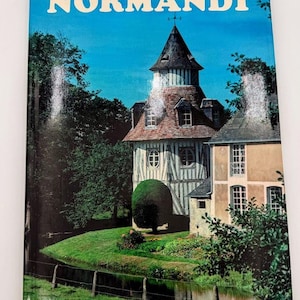 May include: A hardcover book titled NORMANDY featuring a color photograph of a half-timbered stone building with a conical roof, a tall tower, and multiple windows, situated next to a small stream with green grass and trees under a blue sky.