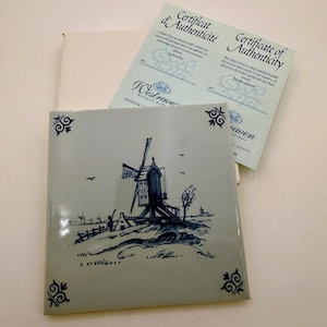 Può includere: Una piastrella quadrata in ceramica con una scena di mulino a vento dipinta a mano in blu su sfondo grigio chiaro con decorazioni angolari. Include un certificato di autenticità di Westraven Delft con testo in francese e inglese.