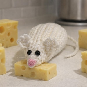 May include: A handmade white knitted mouse with black eyes and a pink nose, perched on a block of yellow cheese. Several other cheese blocks with holes are arranged around the mouse, creating a playful scene.