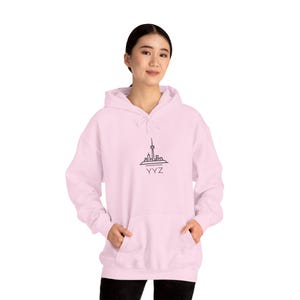 May include: Light pink hooded sweatshirt featuring a black line drawing of a city skyline with the text "YYZ" below it. The sweatshirt has a front pocket and a drawstring hood. The model is wearing the sweatshirt.