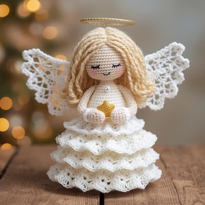 Op de afbeelding: Een handgemaakte gehaakte engelpop met blond haar, een gouden halo en witte kanten vleugels. De engel draagt een witte jurk en houdt een gouden ster vast. De achtergrond toont een vage kerstboom, wat een feestelijk thema suggereert.