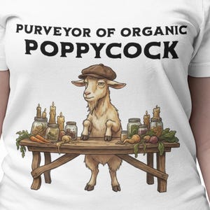 May include: White t-shirt with the text "PURVEYOR OF ORGANIC POPPYCOCK". The shirt features a cartoon goat wearing a hat behind a table with jars and vegetables. The person is wearing black trousers and bracelets.