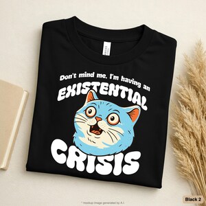 May include: A black t-shirt featuring a graphic of a wide-eyed light blue cat with orange inner ears. The text above the cat reads "Don't mind me, I'm having an" and the text below the cat reads "EXISTENTIAL CRISIS" in a bold white font.