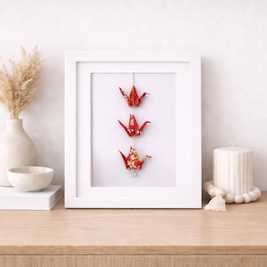 May include: A white framed artwork featuring three origami cranes in shades of red and floral patterns. The artwork is displayed on a wooden surface, alongside a vase with dried flowers, a small bowl, a book, and a candle.