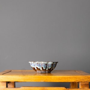 May include: A ceramic bonsai pot featuring a scalloped rim and a reactive glaze that transitions from a deep brown base to a light blue and white drip pattern along the upper edge. The bowl has a wide, shallow shape with visible feet at the bottom.