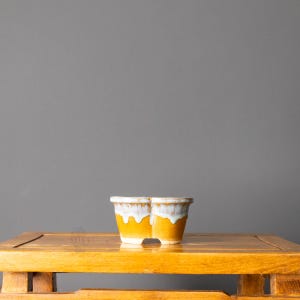 May include: A small, oval-shaped ceramic planter featuring a vibrant mustard yellow base and a contrasting white glaze that drips down from the rim. The planter has a wide, flared opening and a sturdy, thick-walled construction with a smooth finish.