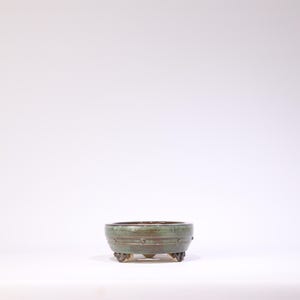 May include: A small, round, green ceramic bonsai pot with three decorative feet. The pot has a dark brown interior and a textured exterior with horizontal bands and small circular details. The pot is designed for bonsai trees.