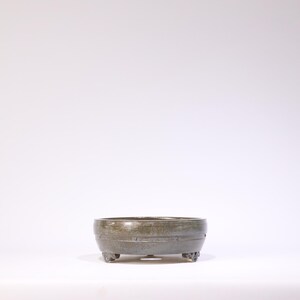 May include: A small, round, gray-green ceramic bonsai pot with a textured surface. The pot has a low profile, decorative bands, and is supported by three small, ornate feet. The pot is designed for planting small trees or plants.