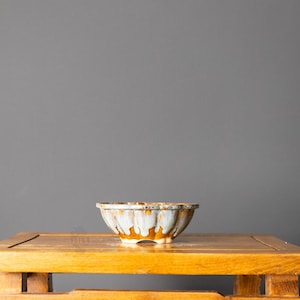 May include: A shallow, round ceramic bowl featuring a fluted, scalloped edge design. The exterior glaze displays a layered aesthetic with a warm, earthy brown base color partially covered by a flowing, creamy white drip pattern throughout.