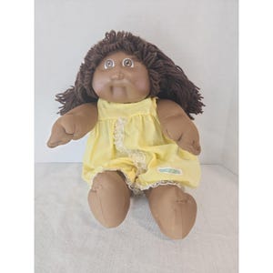 Könnte beinhalten: Eine Stoffpuppe mit dunkelbraunen Wollhaaren und brauner Haut. Die Puppe trägt ein gelbes ärmelloses Kleid mit einem dezenten Karomuster, einer weißen Spitzenborte, die diagonal über die Vorderseite verläuft, und einem kleinen rechteckigen Etikett.