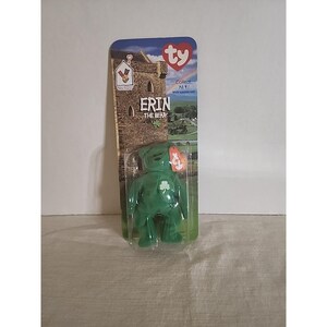 May include: A green plush bear toy named Erin the Bear, packaged in a clear plastic blister on a cardboard backing. The backing features a stone castle, a green landscape, a rainbow, and the text Erin the Bear and Ty logo at the top.