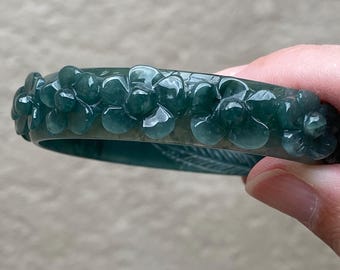 May include: A dark green translucent bangle bracelet featuring a carved floral pattern along the outer surface. The bracelet has a smooth, polished finish with raised flower motifs that wrap around the circumference of the circular band.