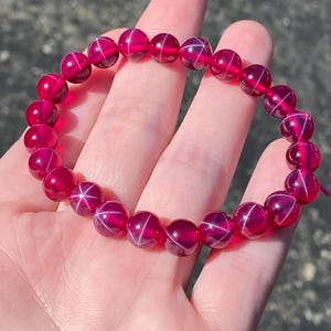 May include: A striking red beaded bracelet featuring a starburst design. The bracelet is composed of round, polished beads, each showcasing a white star pattern. The beads are strung together, creating a circular form. The bracelet is held in a hand.