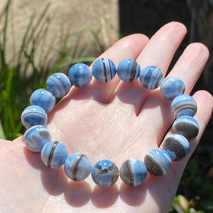 May include: A stretch bracelet composed of round, polished beads in shades of light blue and white, featuring distinct dark brown or black horizontal banding patterns throughout the stones, arranged in a circular loop on an elastic cord.