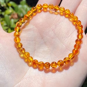 May include: A circular bracelet composed of small, polished, translucent amber beads in varying shades of golden yellow and deep orange. The beads are strung together on an elastic cord, creating a continuous, flexible loop of smooth, rounded stones.