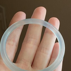 May include: A smooth, translucent, pale blue-grey bangle bracelet with a polished, rounded surface. The circular accessory features a consistent thickness and a clear, glass-like appearance that allows light to pass through its solid, continuous band.