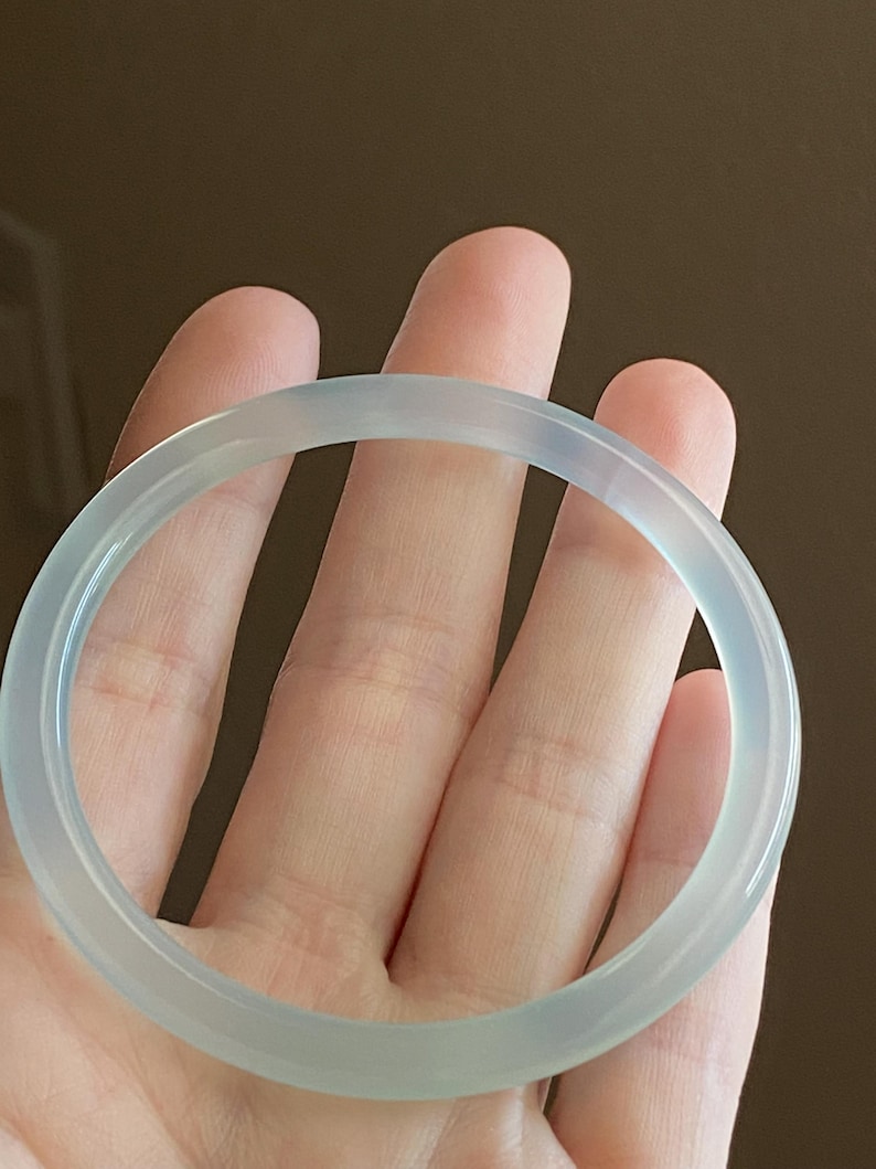 May include: A smooth, translucent, pale blue-grey bangle bracelet with a polished, rounded surface. The circular accessory features a consistent thickness and a clear, glass-like appearance that allows light to pass through its solid, continuous band.
