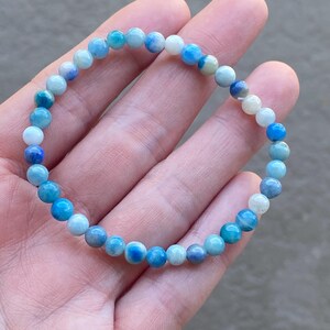 May include: A beaded bracelet featuring a variety of blue and white round beads. The bracelet is made of small, polished stones in shades of light blue, dark blue, and white. The bracelet is shown on a hand.