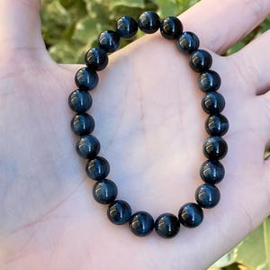 May include: A stretch bracelet composed of round, polished dark blue tiger eye beads. The beads exhibit a chatoyant lustre with deep navy and charcoal tones, arranged in a continuous circular pattern on an elastic cord to form the wrist accessory.