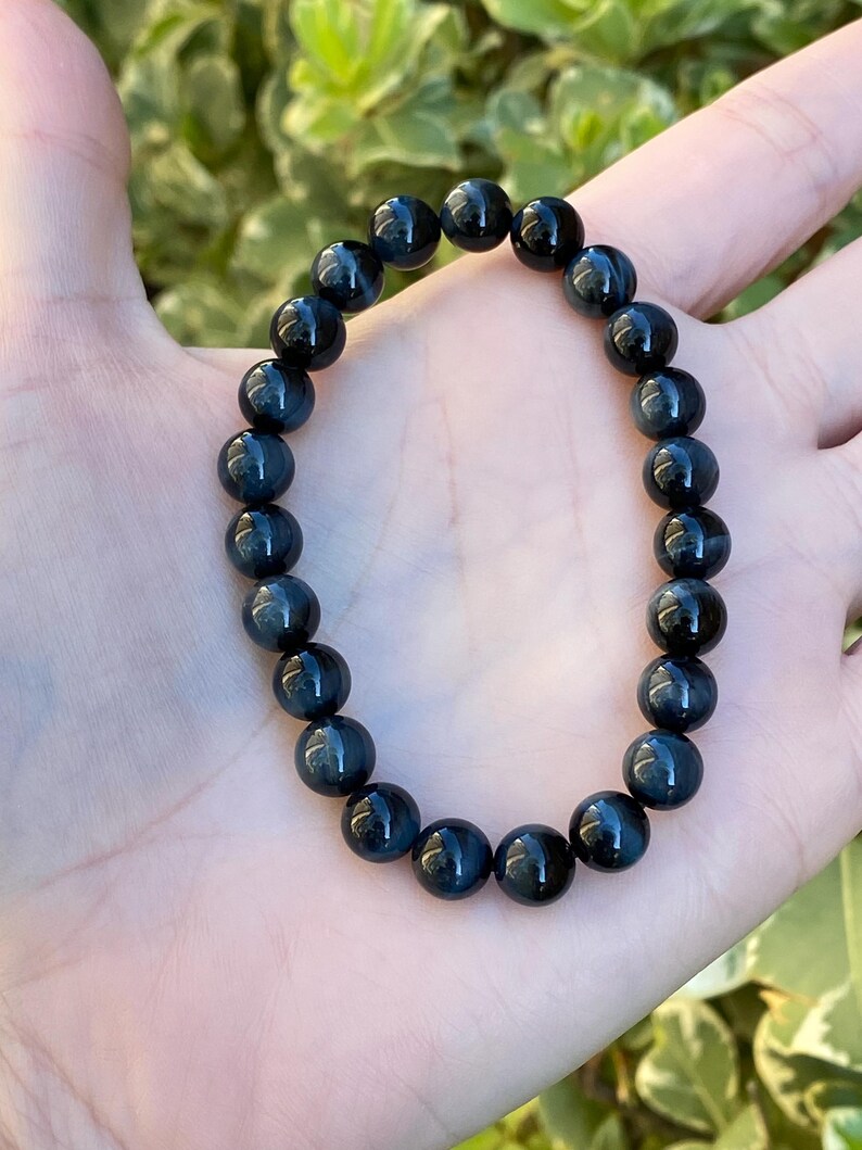 May include: A stretch bracelet composed of round, polished dark blue tiger eye beads. The beads exhibit a chatoyant lustre with deep navy and charcoal tones, arranged in a continuous circular pattern on an elastic cord to form the wrist accessory.