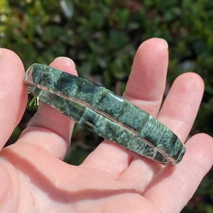 May include: A segmented bracelet composed of rectangular, dark green, mottled stone beads. The beads feature a polished, glossy finish with visible fibrous, feathery inclusions throughout the deep forest green surface of each individual segment.