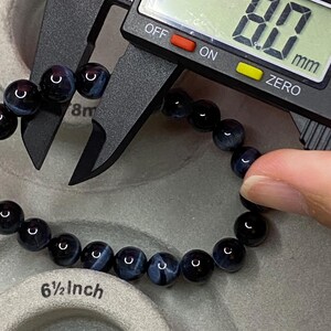 May include: A bracelet made of round, dark blue and black tiger eye beads. A black digital caliper measures one bead at 8.0 mm. The bracelet is positioned near a grey circular display stand with the text 6 1/2 inch printed on its surface.