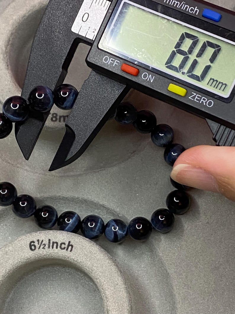 May include: A bracelet made of round, dark blue and black tiger eye beads. A black digital caliper measures one bead at 8.0 mm. The bracelet is positioned near a grey circular display stand with the text 6 1/2 inch printed on its surface.
