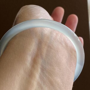 May include: A smooth, translucent white bangle bracelet with a rounded profile. The material appears to have a soft, milky quality with subtle light diffusion throughout the circular band, creating a clean and simple aesthetic for the wrist.