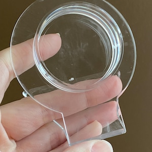 以下が含まれることがあります： 透明な円形のプラスチック製のオブジェクト。平らなベースと、隆起した同心円のデザインが特徴です。手で持たれており、その透明性と形状が示されています。装飾的または機能的なアイテムを示唆するデザインです。