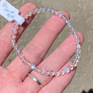 May include: A clear beaded bracelet with iridescent beads. The bracelet is held in a hand, and a tag with the text "BL Moon 3/14" is attached. The bracelet is made of small, round beads.